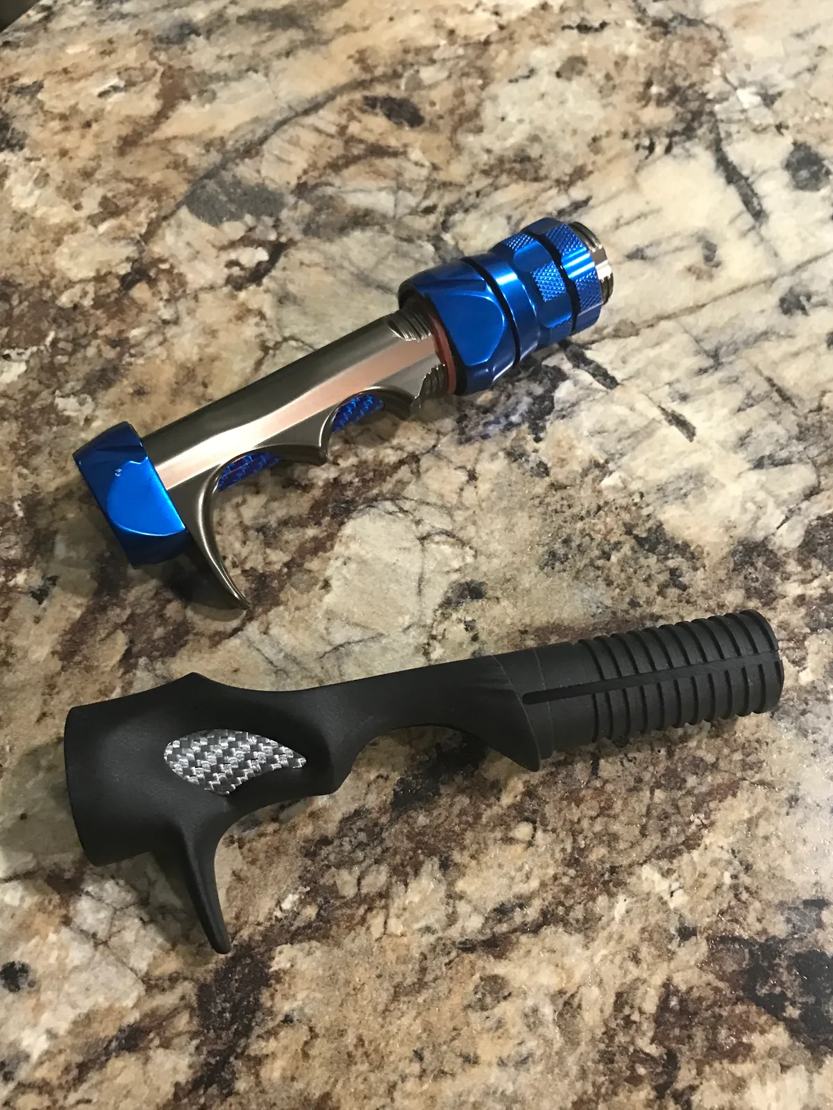 Two casting-rod handles lying on a stone counter, each with a curved finger trigger under the reel seat; the upper one has a threaded barrel and knurled locking nut.