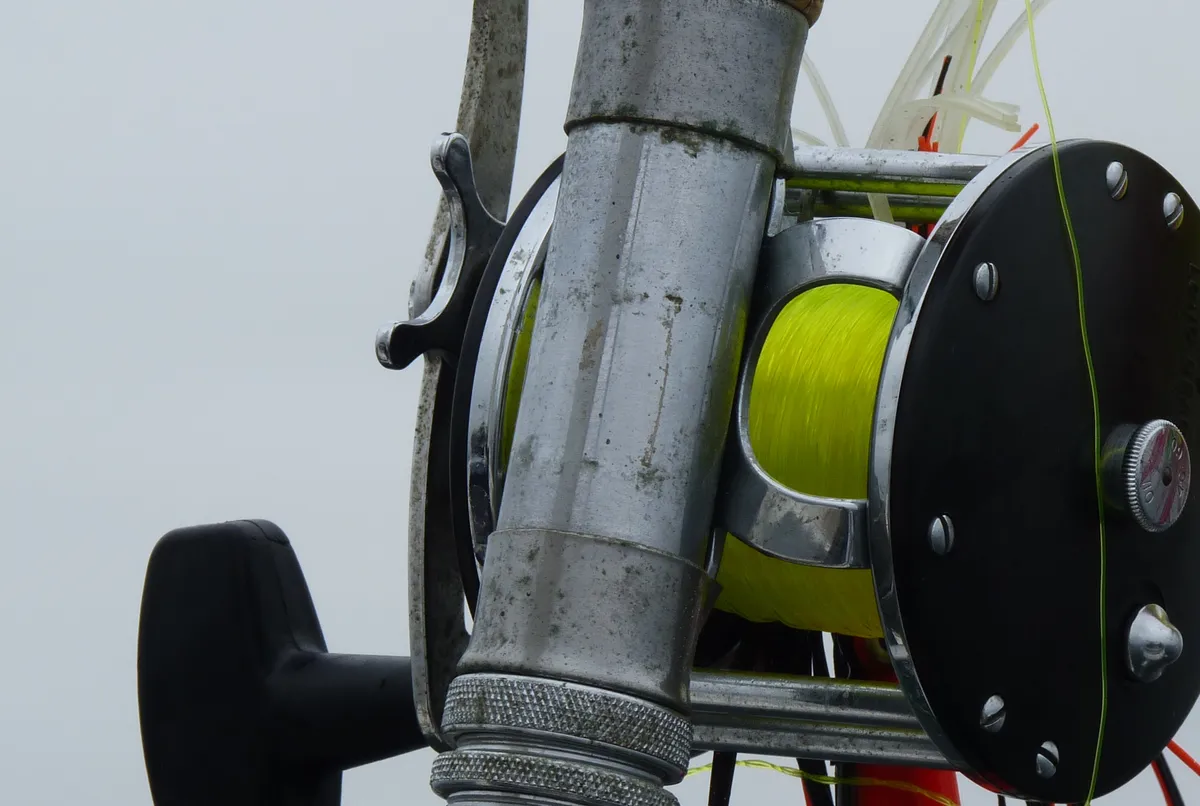 A round revolving-spool reel clamped to a rod, with a T-shaped handle, a star-shaped wheel beside it, a spool of yellow line between two side plates and knurled locking rings on the reel seat.