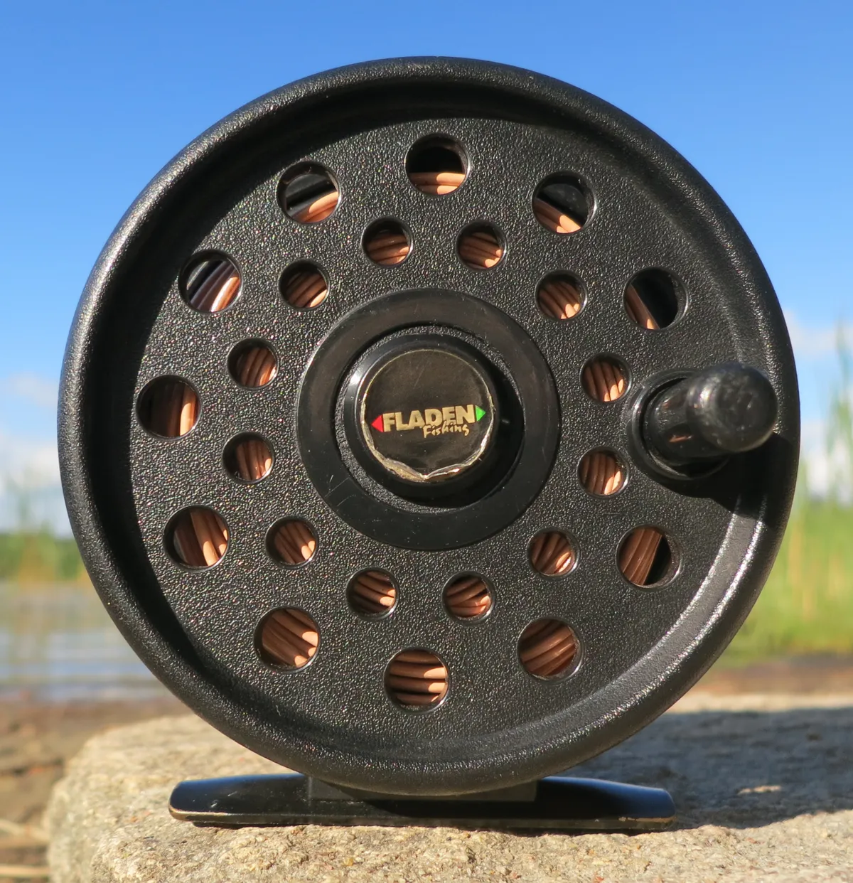 A black fly reel seen face-on, standing on its foot, with coiled fly line showing through holes in the spool face and a small handle knob near the rim.