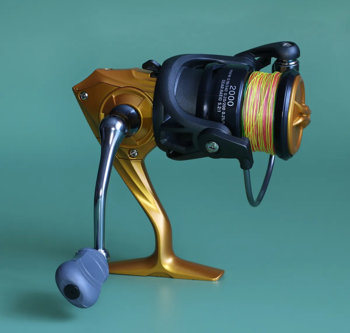 Spinning reel photographed off the rod, with its spool, curved bail wire and handle visible against a teal background.